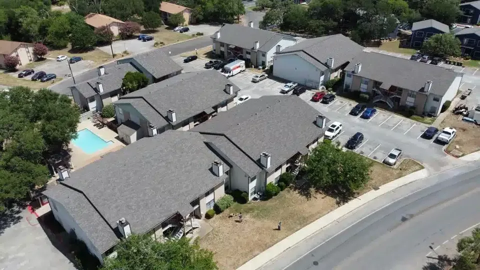 Aerial view of a multi-building apartment community with gray asphalt shingle roofs, parking areas and a pool